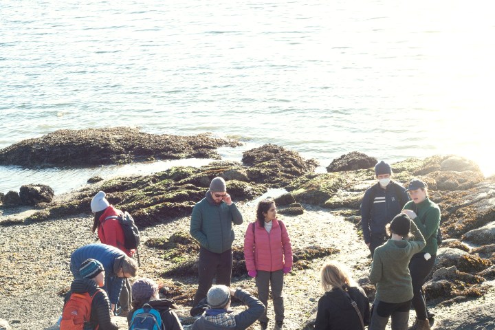 Group of people in winter clothing standing on a rocky shore by the water.