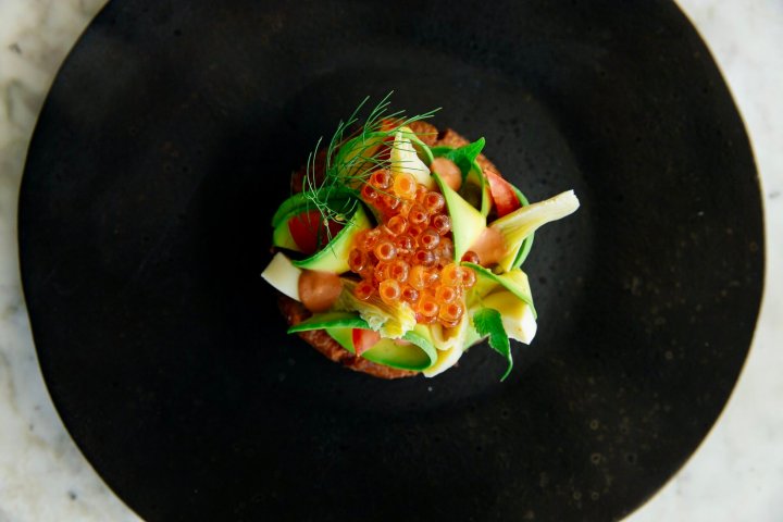 Gourmet dish with avocado slices, orange roe, herbs on a dark plate.